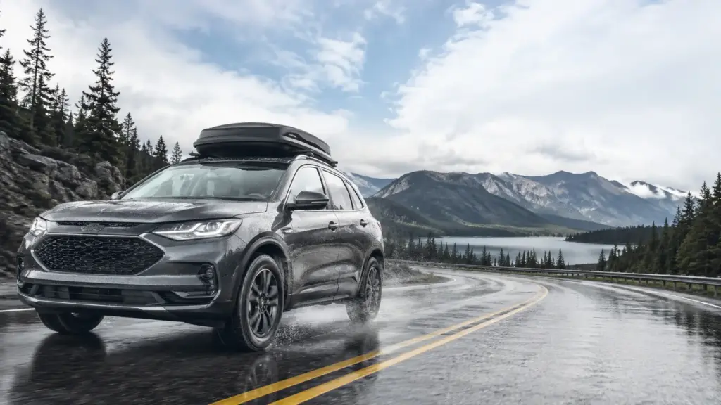 Silver/gray SUV with a rooftop cargo box on a wet, winding mountain road beside a lake and pine forest, during cloudy weather.